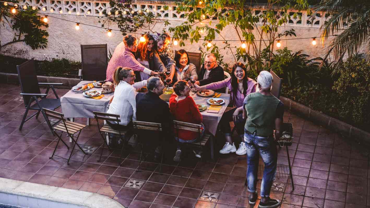 A large family eats outdoors by the pool under the glow of string lights.