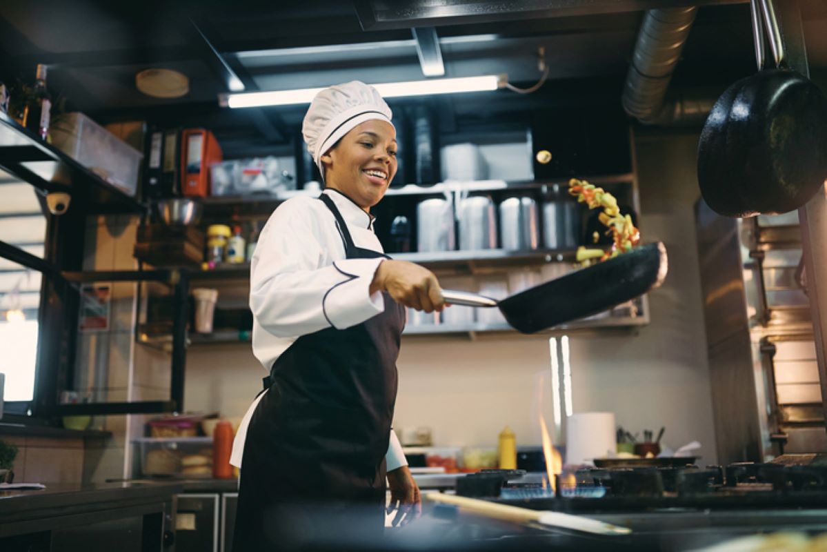 A happy chef preparing food in a frying pan behind a range